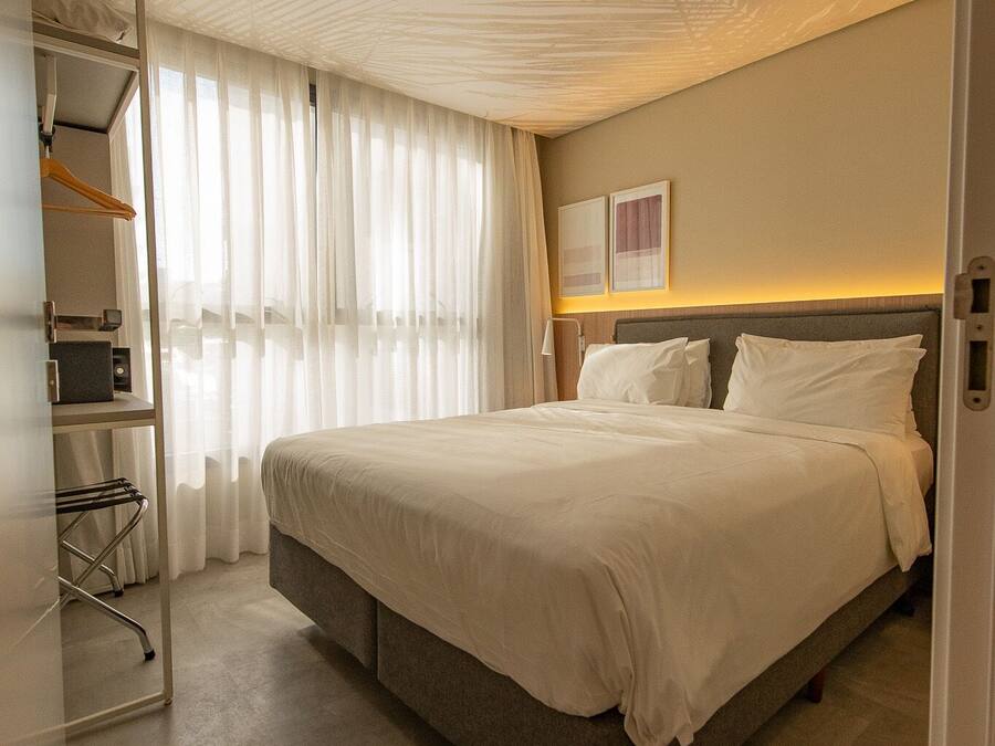 Habitación de hotel con una cama grande, un armario, un espejo y una ventana con cortinas translúcidas.