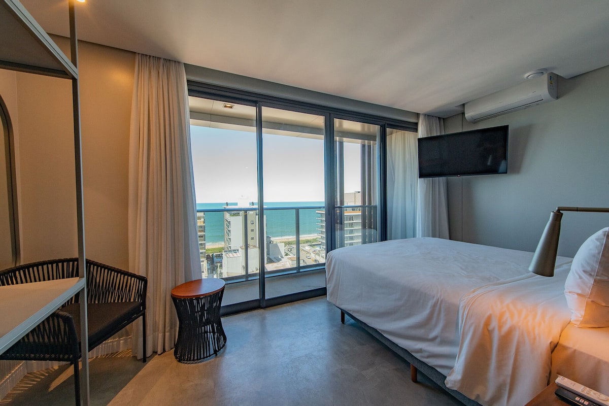 A hotel room with a large window, a bed, a TV, and a small table.