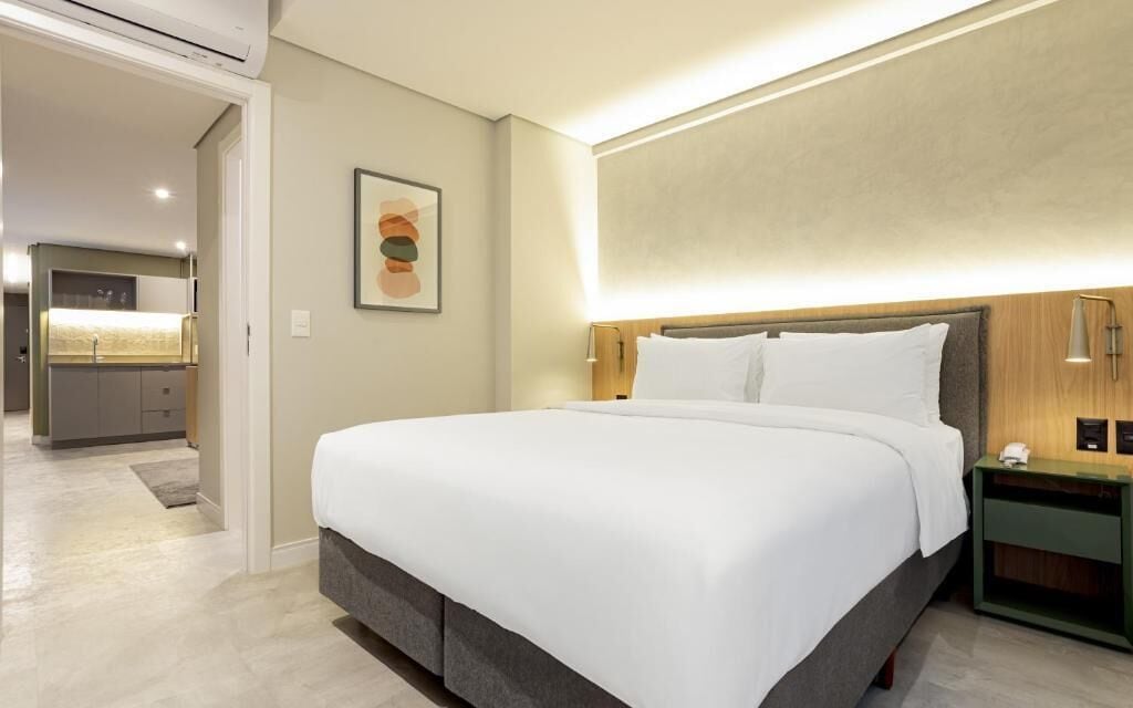 A modern hotel room with a large bed, a bedside table, a wall-mounted light, and a framed picture on the wall.