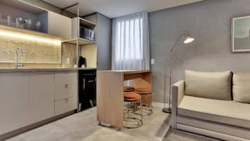 A modern kitchen with a built-in oven, a dining area with a sofa and a lamp, and a large window with curtains.