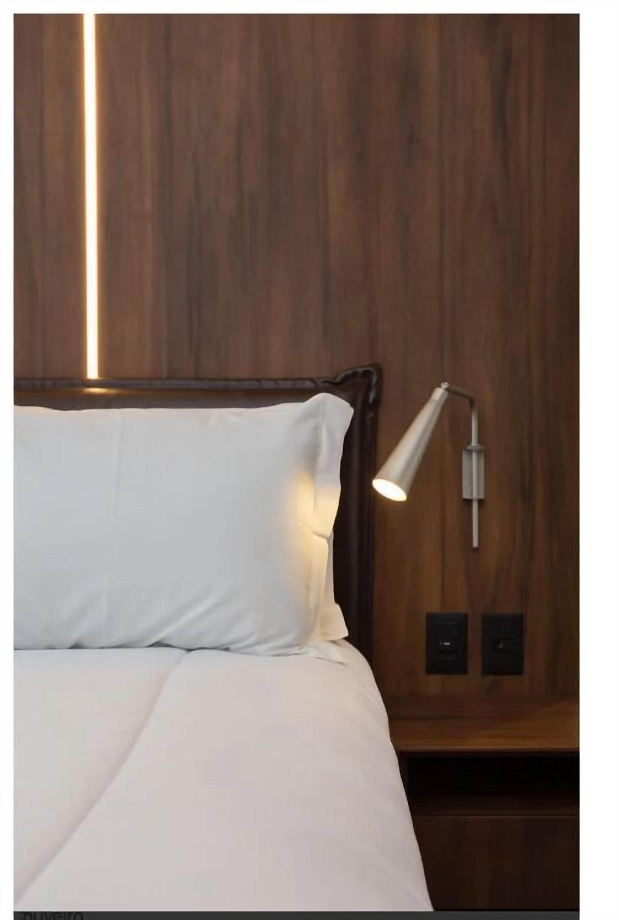 A bed with white bedding, a wooden headboard, and a wall-mounted nightstand with a lamp.