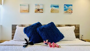 A neatly made bed with white bedding and two blue pillows, a pink orchid on the bed, and four beach-themed paintings on the wall. - Tu Paraíso Tropical, Terraza y Piscina Soleada, Bañera con Hidromasaje y Jardin (Noord)