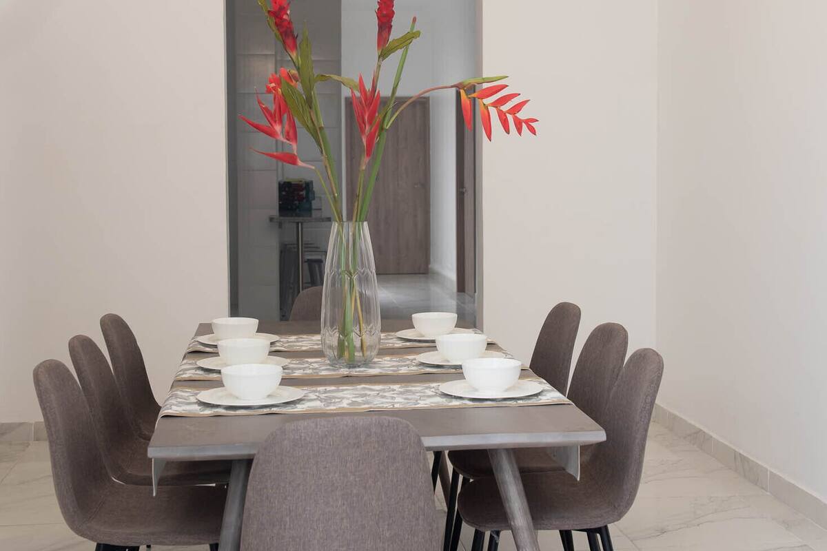 A dining area with a rectangular table set for six, surrounded by six chairs. A vase with red flowers is placed on the table.