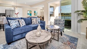 A modern living room with a blue sofa, a marble coffee table, and a patterned armchair. A large window offers a view of a body of water.