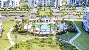 A modern hotel complex with a central swimming pool surrounded by palm trees and lounge chairs.