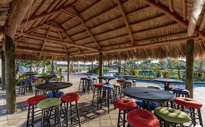 Un patio al aire libre con techo de paja, mesas redondas y taburetes de colores.
