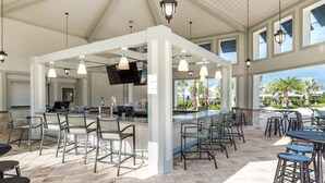 A covered outdoor bar area with multiple seating options, including bar stools and chairs, and a flat-screen TV mounted on the wall.