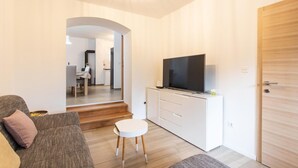 Un saló modern amb un sofà, una taula rodona petita i una televisió sobre un moble blanc. Hi ha una porta de fusta i un passadís que condueix a una zona de cuina.
