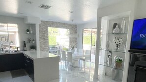 A modern kitchen with a marble countertop, a dining area with white chairs, and a TV on the right. - JC’s Modern Paradise (Chula Vista)