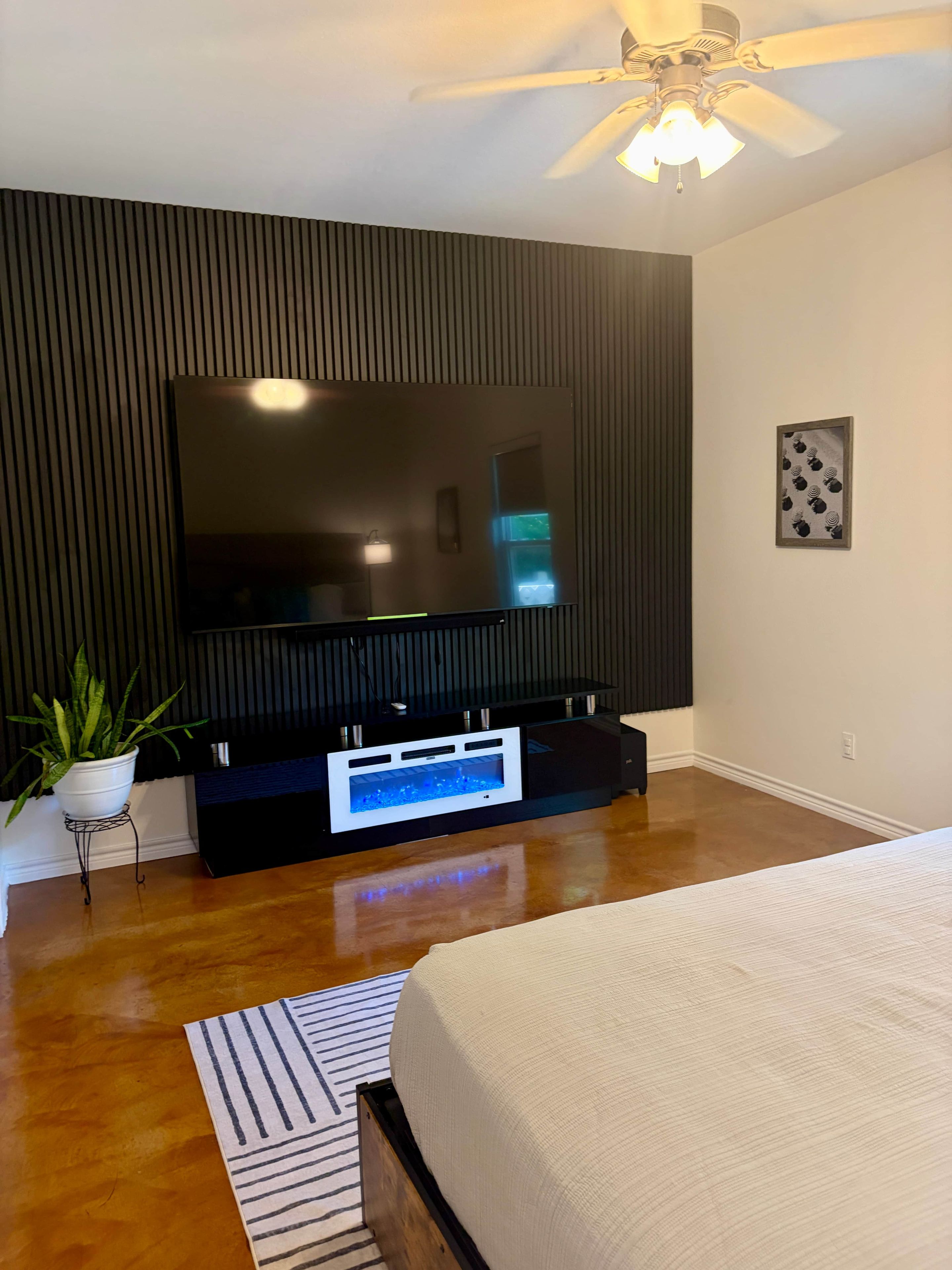 Ein modernes Schlafzimmer mit einem großen Flachbildfernseher an einer dunklen Wand, einem Deckenventilator, einer Topfpflanze und einem Bett mit einem Streifen Teppich.
