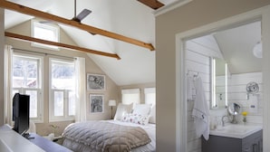 A bedroom with a bed, a desk, a chair, a window, and a bathroom with a sink and mirror.