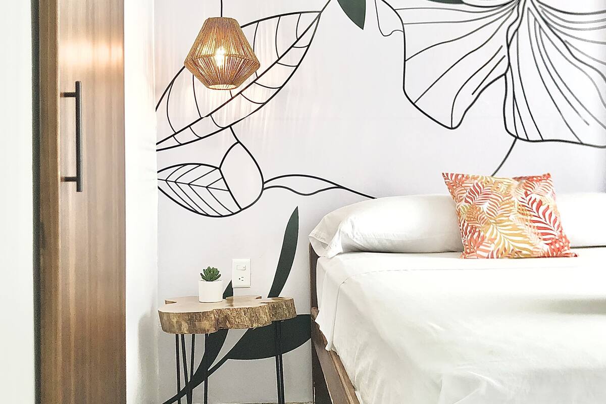 Une chambre à coucher avec un lit, une table de chevet, une petite table avec une plante et un mur orné d’un motif de feuilles.