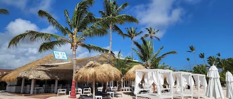 A beachside restaurant with thatched roofs, white umbrellas, and lounge chairs.