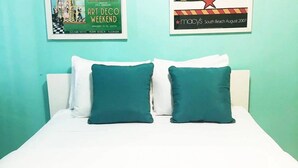 A bed with white bedding and two teal pillows, a framed poster of the Art Deco Weekend, and a Macy's sign on the wall. - Royal Hotel Unit 337 (Miami Beach)