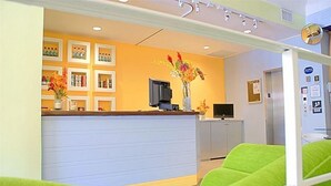 A reception area with a counter, a television, and a vase of flowers. There are green chairs in the foreground. - Royal Hotel Unit 337 (Miami Beach)