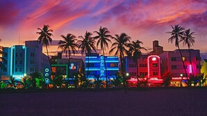 A row of brightly lit hotels with neon signs, palm trees, and a sunset sky. - Royal Hotel Unit 337 (Miami Beach)