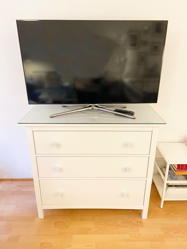 Un mueble blanco para televisor con un televisor de pantalla plana, dos controles remotos y un estante pequeño blanco con libros.