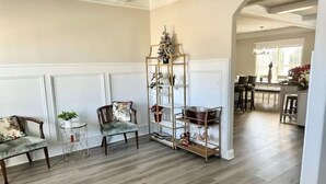 A living room with a wooden floor, two upholstered chairs, a tall metal shelf with decorative items, and a dining area in the background. - Spacious & Cozy Turnkey Master’s Tournament Rental with Concierge Level Service (Grovetown)