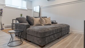 A modern living room with a grey sectional sofa, a small round table, and a large window providing natural light. - Cozy Haven in Port Hope: Explore, Relax, Recharge (Port Hope)