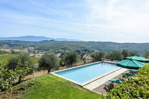 A rectangular pool with a clear blue poolside area, surrounded by green grass and trees, with a view of distant hills and a clear sky.