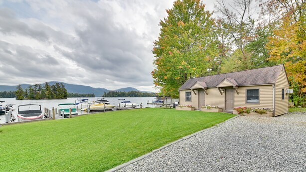 A lakeside cabin with a grassy lawn, a gravel driveway, and boats docked at a pier. - Lake George Waterfront Cabin w/ On-Site Marina! (Bolton Landing)