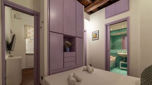 A bedroom with a bed, a small bedside table, a nightstand, a wall-mounted shelf with a vase, and a bathroom visible through an open door.
