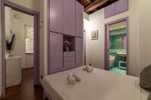 A bedroom with a bed, a small bedside table, a nightstand, a wall-mounted shelf with a vase, and a bathroom visible through an open door.