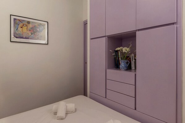 Una habitación con una cama, un armario morado, un cuadro abstracto enmarcado y un jarrón con flores sobre el armario.