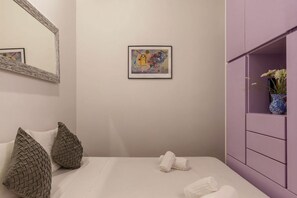 A bedroom with a bed, pillows, a framed artwork, a mirror, and a purple cabinet with a vase.