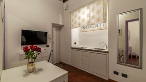 A kitchen with a sink, a window with a checkered curtain, a television mounted on the wall, and a vase with red roses on a table.