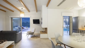 Sala de estar moderna com sofá, mesa de jantar e cadeiras. Há uma TV na parede e uma varanda com vista para o exterior.