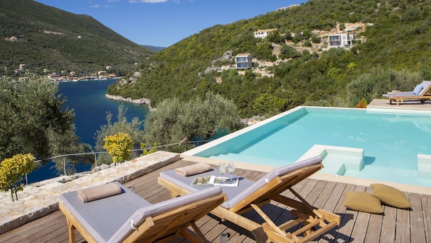Um deck de madeira com espreguiçadeiras e piscina, com vista para uma área costeira com casas numa encosta.