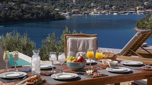 Mesa de jantar posta com pratos, copos e um jarro de água, com vista para um lago com barcos e casas ao fundo.