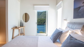 Quarto com cama, mesa de cabeceira, espelho e vista para uma varanda com guarda-corpo e paisagem de montanhas.