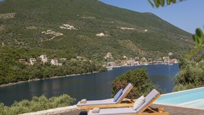 Terraza de madera con sillones de descanso con vista a un lago, rodeada de follaje y edificios.