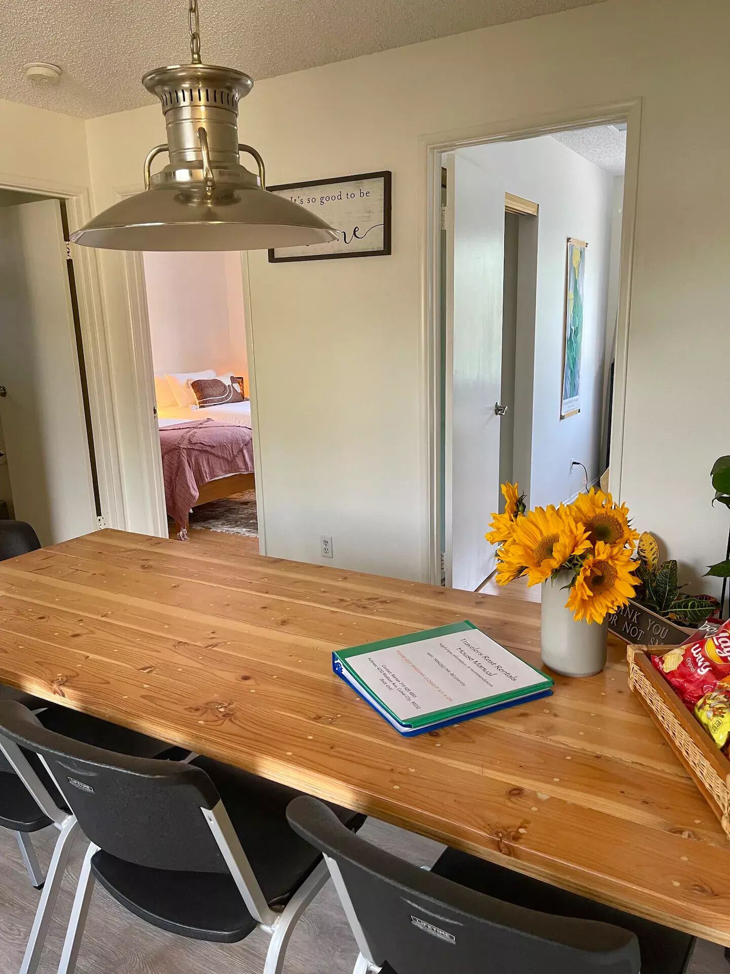 A wooden dining table with chairs, a vase of sunflowers, a framed quote on the wall, and a door leading to a bedroom.
