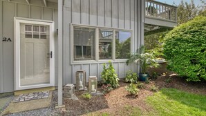 A two-story house with a front door, two windows, and a small garden with potted plants and a wooden deck.