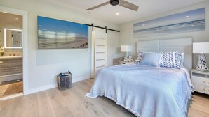 A bedroom with a large bed, a ceiling fan, a painting of a beach, and a view of a bathroom with a mirror.
