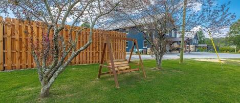 A backyard with a wooden swing set, a fenced area, and a house in the background.