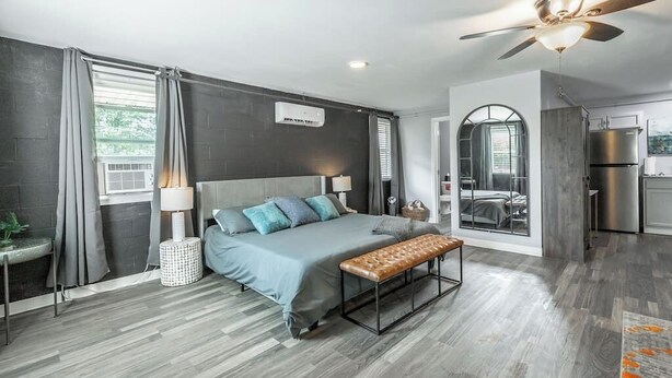 A modern bedroom with a large bed, a bench, a ceiling fan, and a view of the kitchen.