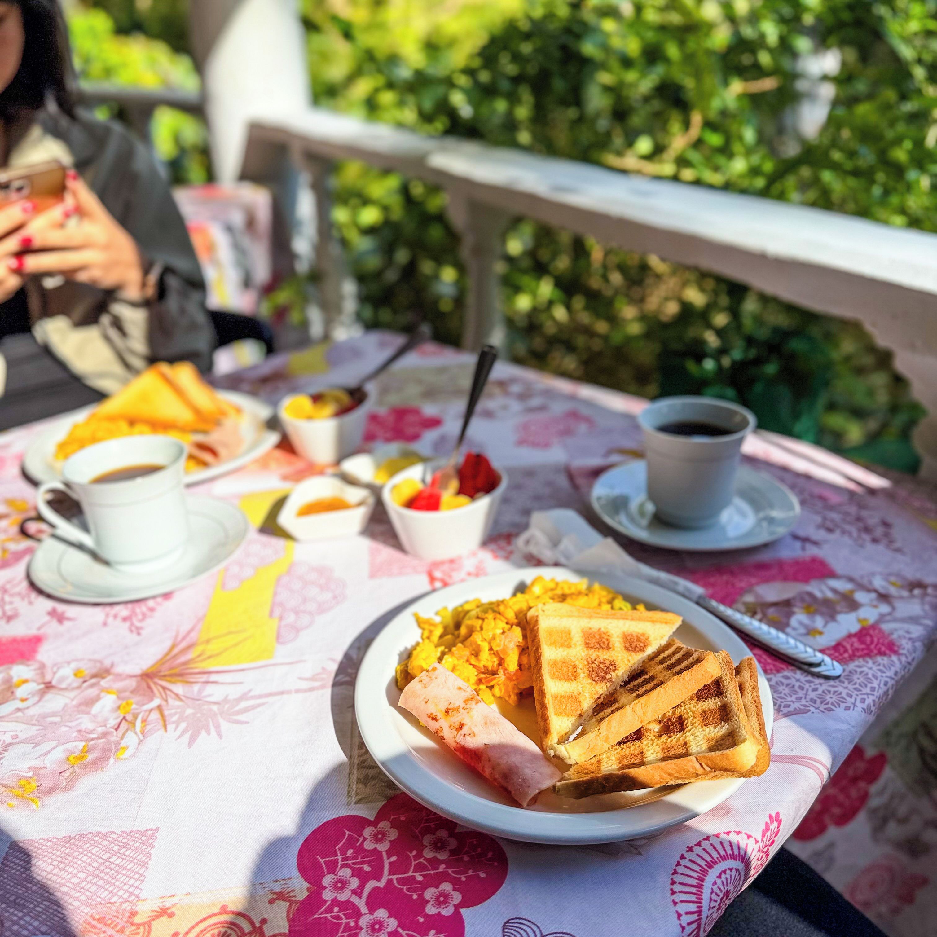 Une table recouverte d’une nappe à motifs floraux, une assiette de nourriture, une tasse de café et une personne tenant un téléphone.