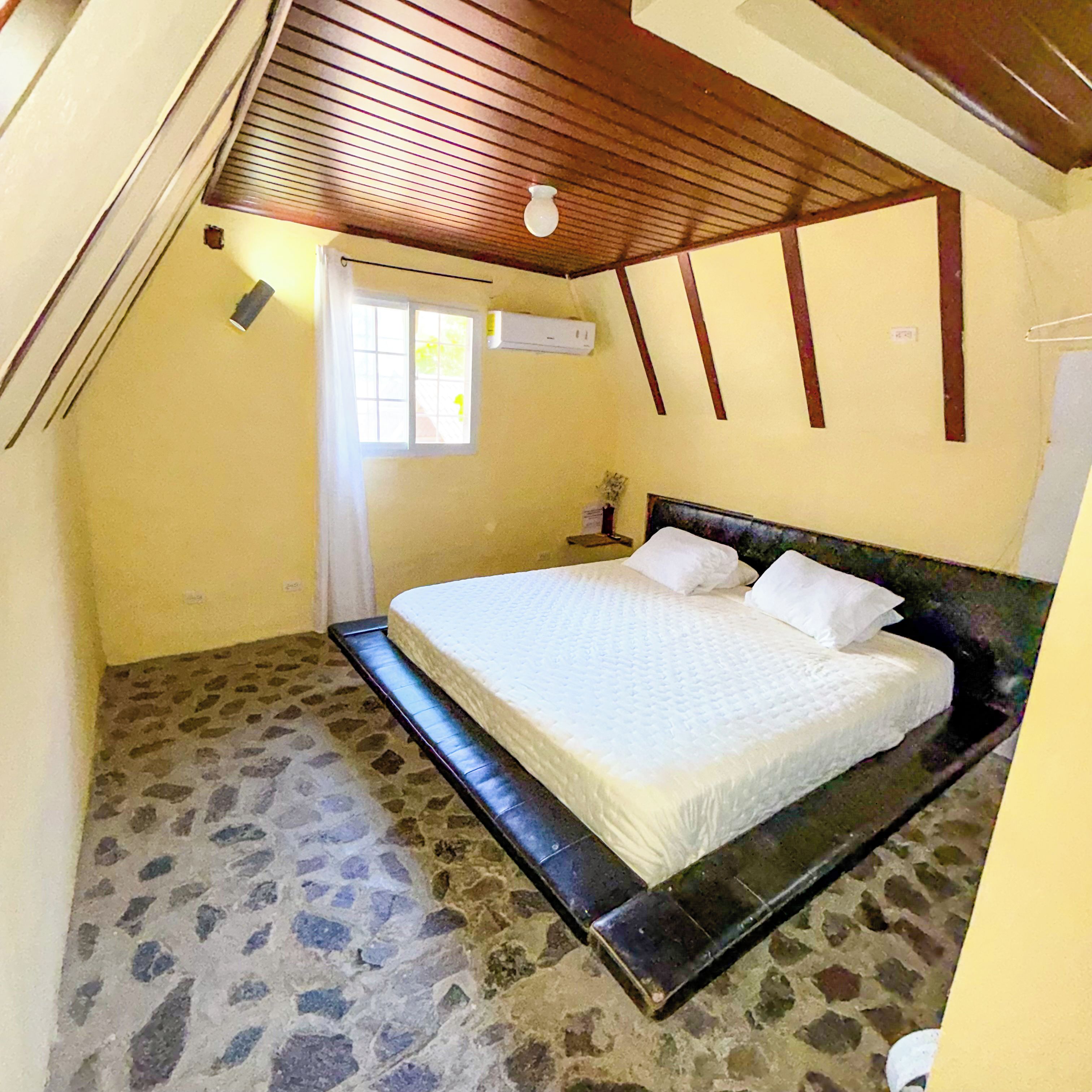 A bedroom with a stone floor, a bed with white bedding, a wooden ceiling, and a window.