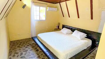 A bedroom with a stone floor, a bed with white bedding, a wooden ceiling, and a window.