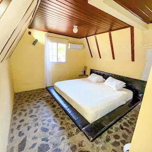 Een slaapkamer met een stenen vloer, een bed met witte beddengoed, een houten plafond en een raam.