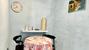 A small room with a round table covered in a floral tablecloth, two chairs, a clock on the wall, a shelf with decorative items, and a painting on the wall. - Refugio Cariguana  (El Valle de Anton)
