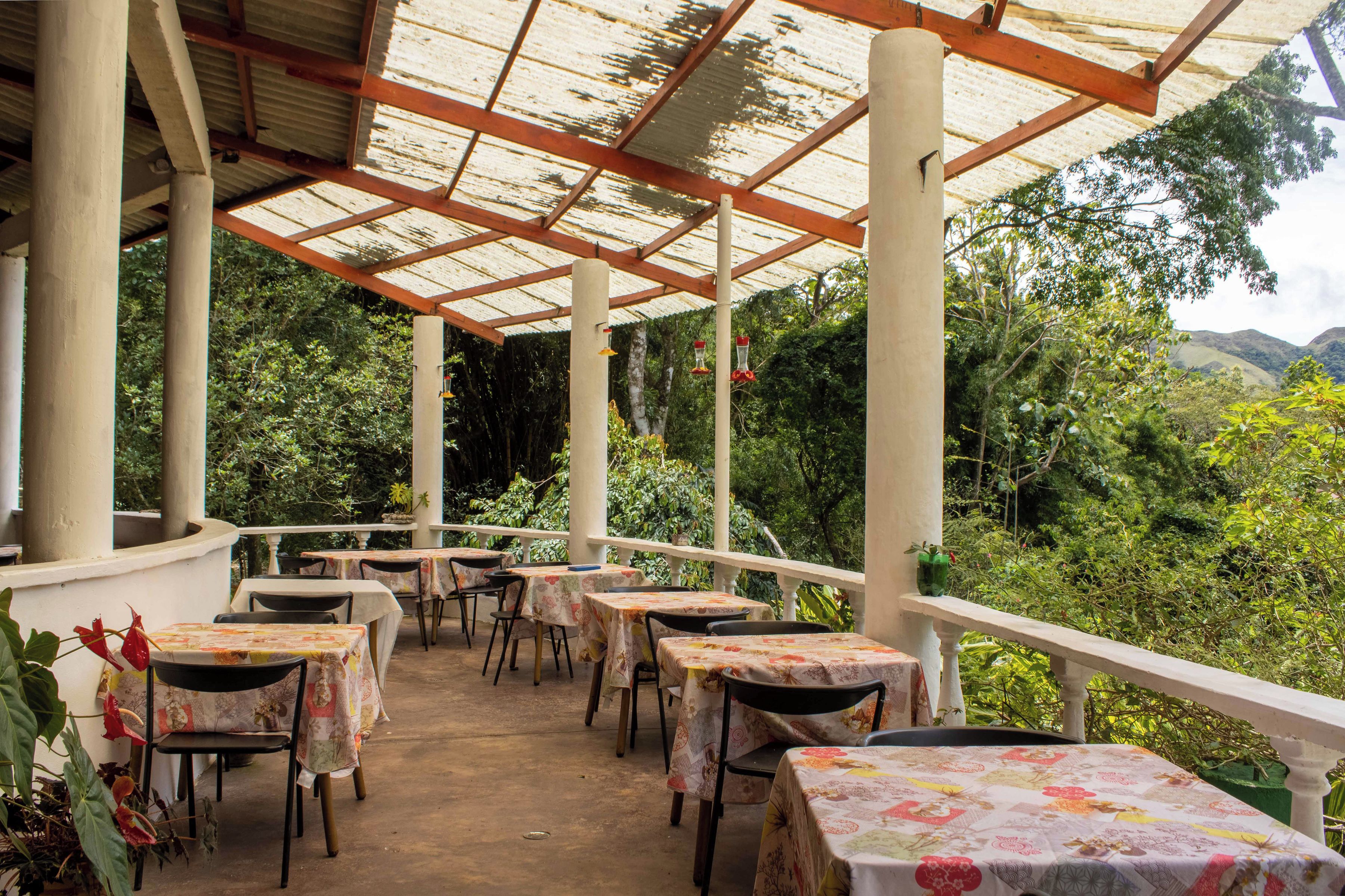 An outdoor covered patio with tables and chairs, surrounded by lush greenery and hills in the background.