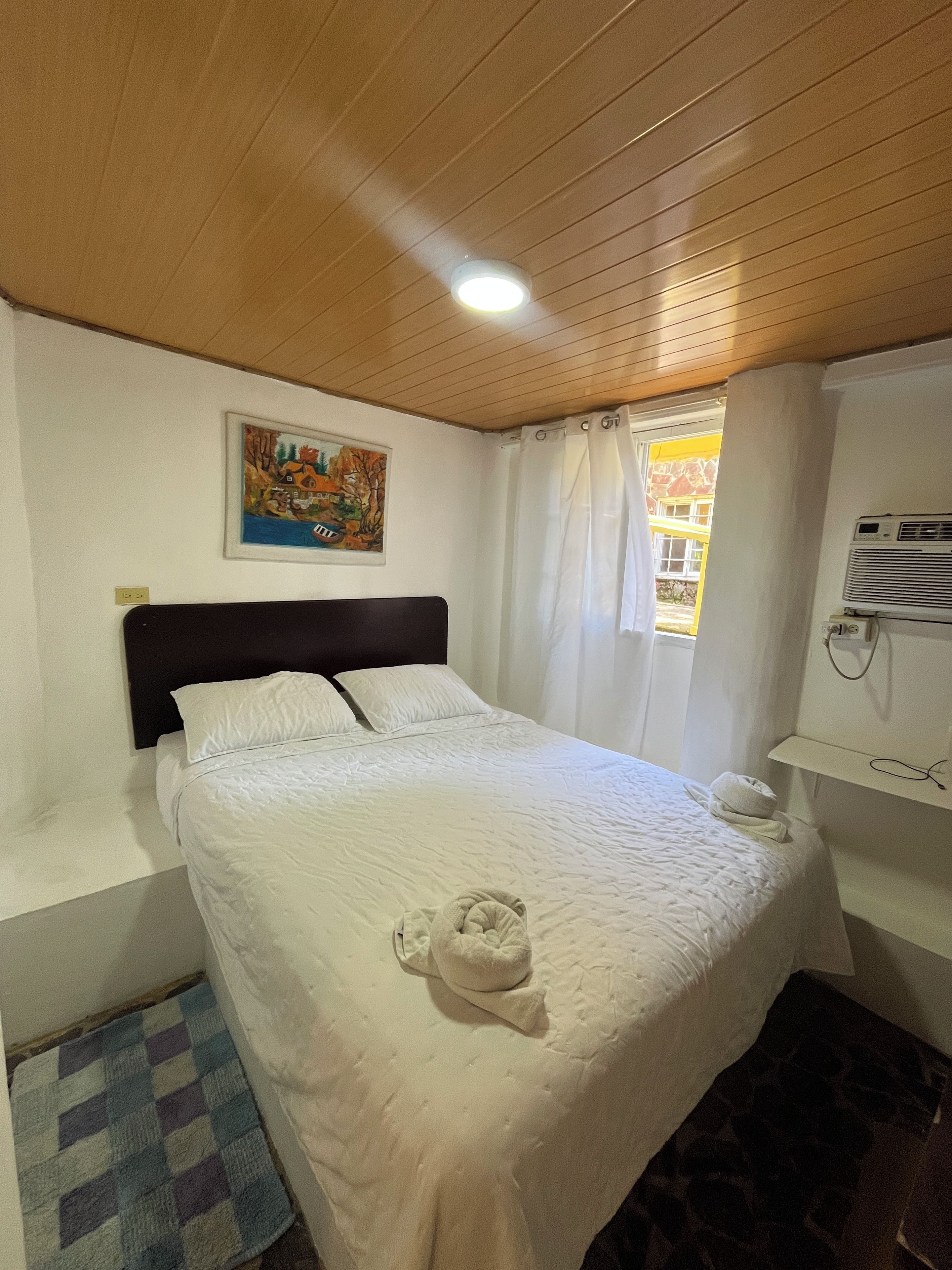 A hotel room with a bed, a painting on the wall, a window with curtains, an air conditioning unit, and a small rug.