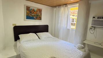 A hotel room with a bed, a painting on the wall, a window with curtains, an air conditioning unit, and a small rug.