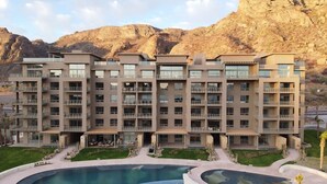 A multi-story hotel with balconies, a swimming pool, and a mountainous backdrop.