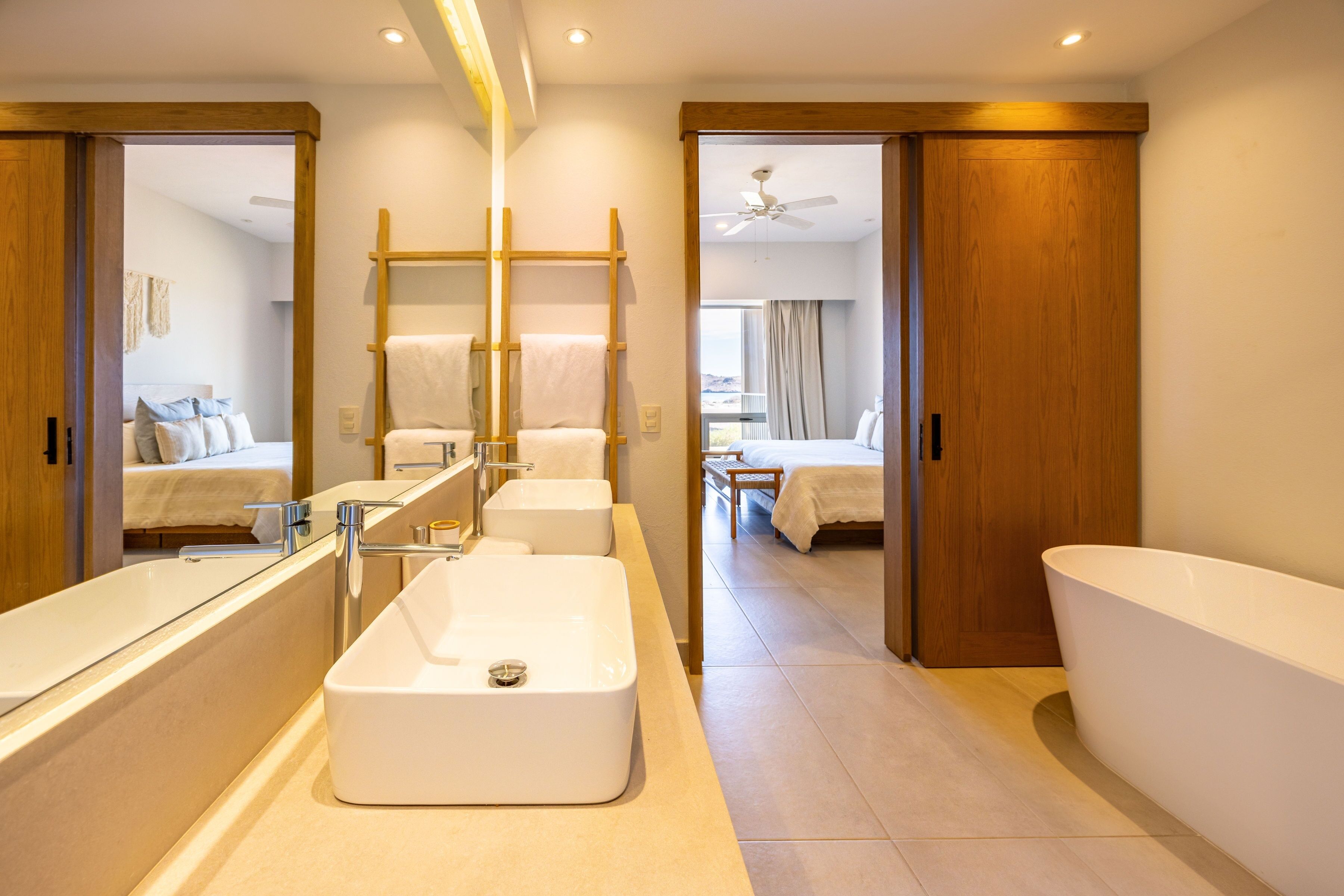 Un baño moderno con una bañera grande, un lavamanos independiente y un espejo. Hay puertas de madera que dan a un dormitorio y un balcón.